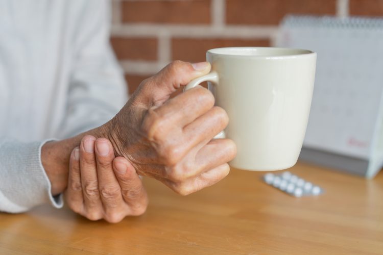 close up senior woman hold on hand to relief shaky symptom while drink water for Parkinson’s disease and chronic illness health care concept