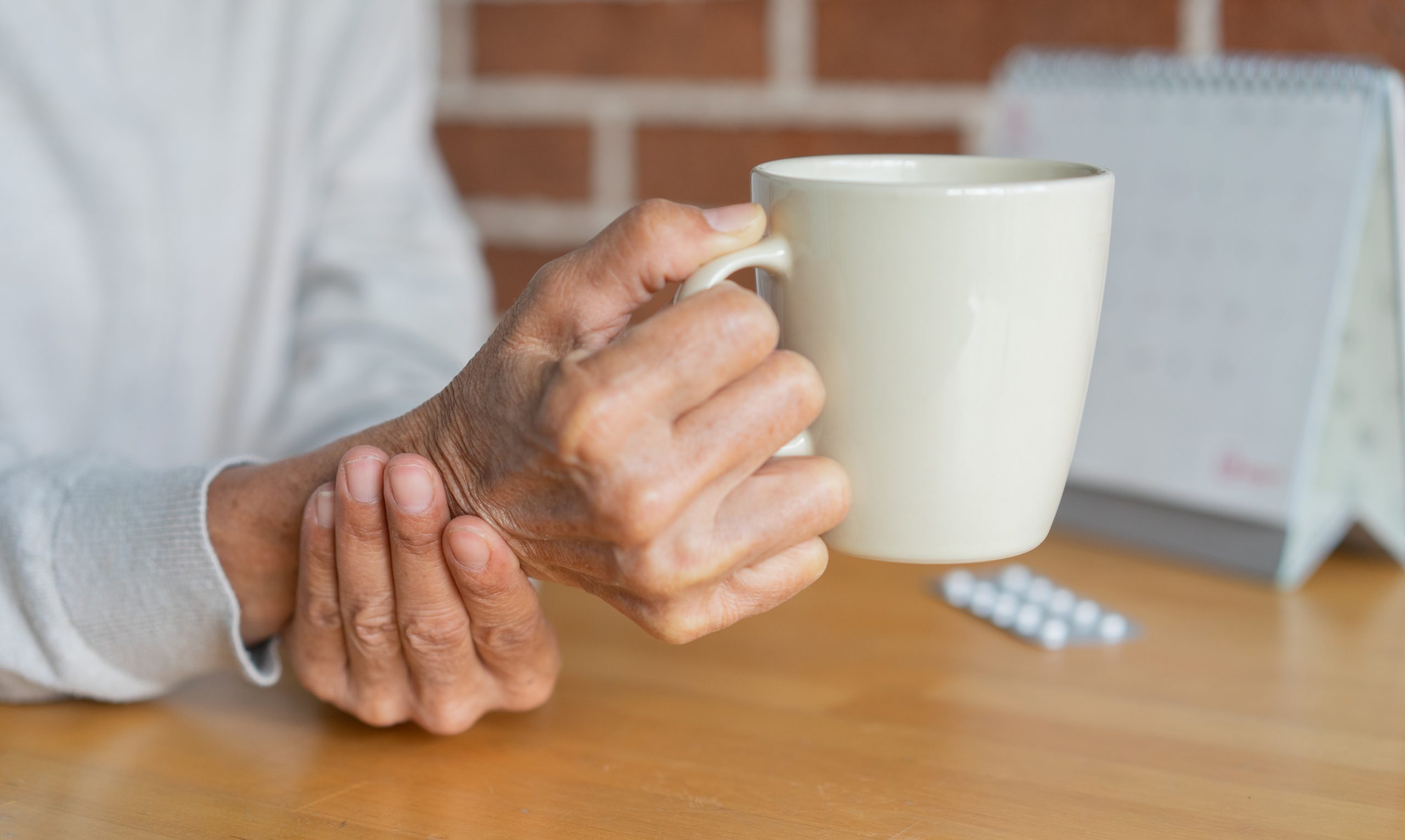 close up senior woman hold on hand to relief shaky symptom while drink water for Parkinson’s disease and chronic illness health care concept